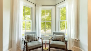 A corner room with two armchairs, a small table, and three windows with white curtains. - Raspberry Inn · Classic Riverfront Home-Mins to Woodstock & Skiing (Bridgewater)