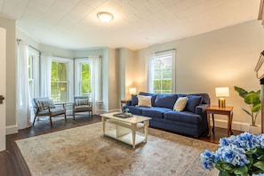 Een woonkamer met een blauwe bank, twee fauteuils, een salontafel en een groot raam met gordijnen.