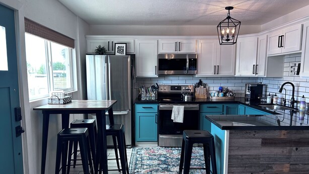 A modern kitchen with white cabinets, a stainless steel refrigerator, a microwave, and a sink. There are two bar stools and a dining table. - Modern Farmhouse in Midtown with fully fenced yard (Tucson)
