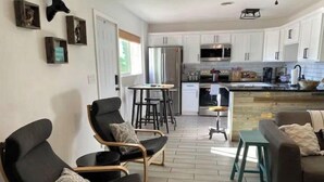 A modern kitchen with white cabinets, stainless steel appliances, and a bar area. There are two black leather chairs with patterned cushions. - Modern Farmhouse in Midtown with fully fenced yard (Tucson)