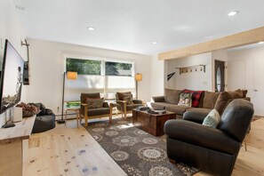 A living room with a sofa, armchair, coffee table, and a rug. There is a window with blinds, a lamp, and a coat rack.