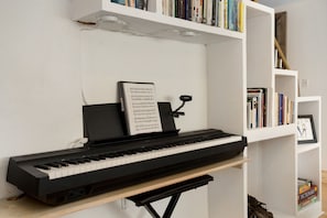 A piano with sheet music on a stand, a bookshelf with books, and a framed picture on a shelf.