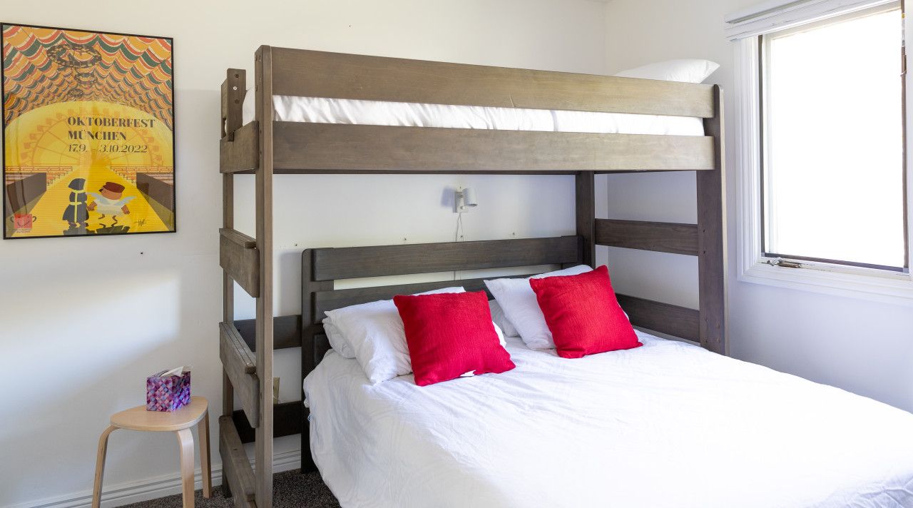 Una cama litera con ropa blanca y almohadas rojas, un taburete de madera y un cartel en la pared.
