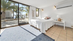 A bedroom with a large sliding glass door leading to a balcony, a bed with a white bedspread, a small bedside table with a lamp, and a view of palm trees and a building outside. - Beachfront Villa + Casitas w/ pool. 6 BR (Los Barriles)