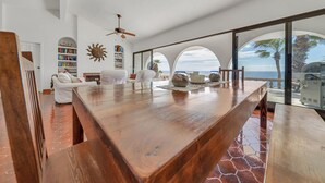 A spacious living area with a wooden coffee table, white sofas, a bookshelf, and large windows offering a view of the ocean. - Beachfront Villa + Casitas w/ pool. 6 BR (Los Barriles)