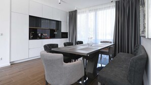 A modern kitchen with white cabinets, a black countertop, and a built-in oven. There is a dining area with a table and chairs, and a large window with curtains.