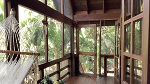 Eine Holzterrasse mit einer Hängematte, einem Tisch mit Obstschale und großen Fenstern mit Blick auf einen tropischen Garten.