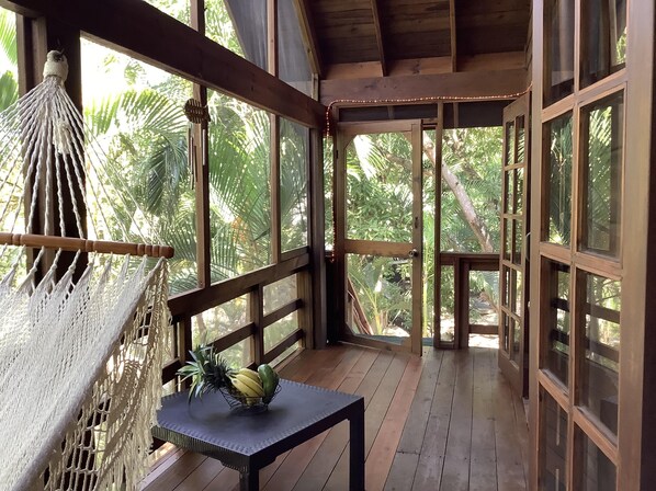 A wooden deck with a hammock, a table with a fruit bowl, and large windows overlooking a tropical garden.