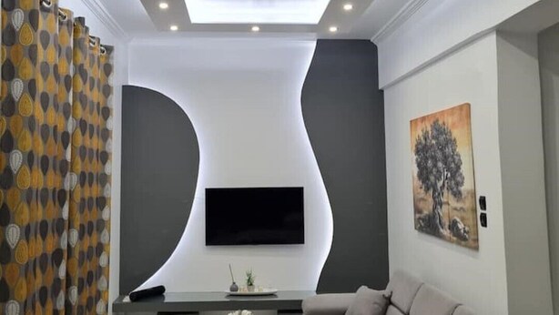 A modern living room with a sofa, a coffee table, and a flat-screen TV mounted on a wall with a unique design. - Acropolis Lux 2 Bedroom Apt,CBD, H/TUB, fast Wifi-1min from Museum&Metro station (Athens)