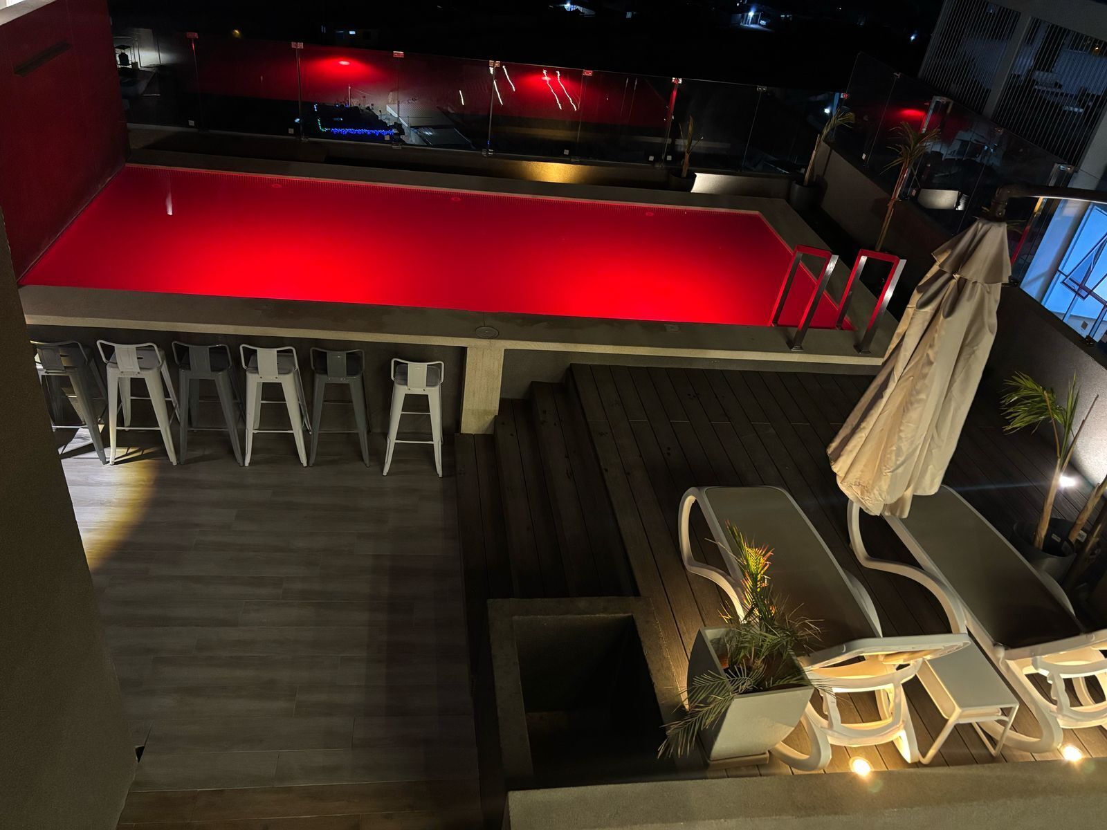Zona de piscina con piscina iluminada en rojo, sillas reclinables blancas y terraza de madera.