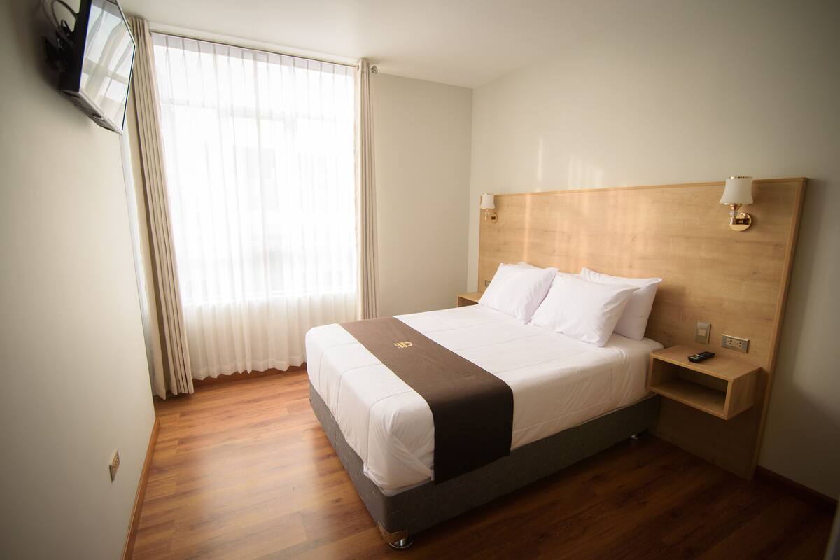 Ein Hotelzimmer mit massivem Holzkopfende, einem Bett mit weißer Bettwäsche, einem Nachttisch mit Lampe und einem an der Wand befestigten Fernseher.