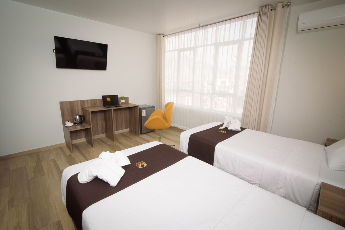 A hotel room with two beds, a desk with a laptop, a chair, and a TV on the wall.
