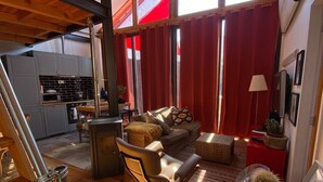 Uma sala espaçosa com piso de madeira, um sofá, uma poltrona e uma mesa de jantar. É possível ver uma cozinha ao fundo, com armários e um micro-ondas.