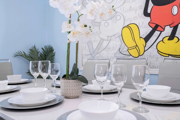 Una mesa de comedor dispuesta con platos, tazones y copas de vino blancos, junto con orquídeas blancas y una planta en maceta.