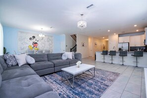 Una sala de estar moderna con un sofá seccional gris, una mesa de centro blanca y un tapete azul y blanco. Hay tres taburetes negros y una cocina con armarios blancos y un refrigerador de acero inoxidable.