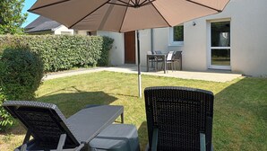 Un espace patio avec un grand parasol, deux chaises longues, une table et une maison en arrière-plan.