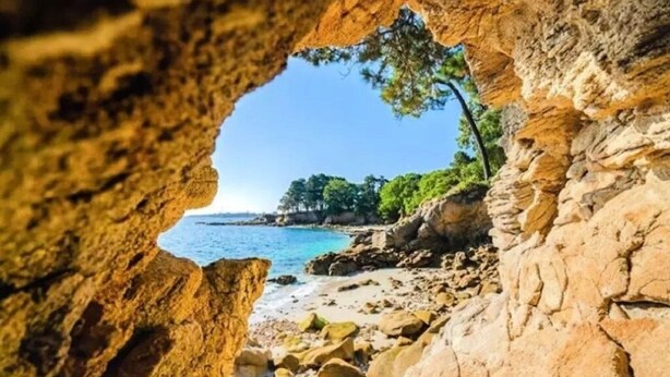 Une vue côtière à travers une arche naturelle formée par des rochers, avec une plage et des arbres en arrière-plan.