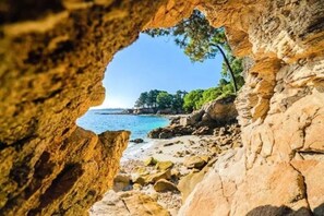 A coastal scene through a natural rock archway with a beach and trees in the background. - 3★ Apartment with Enclosed Garden and Terrace – Peace & Comfort Near the Sea (Fouesnant)