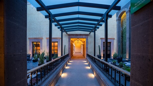 A corridor with a glass ceiling, potted plants, and a sign indicating an evacuation route. - Hotel Patio Santiago (Querétaro)