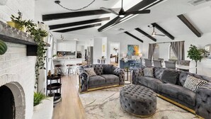 A spacious living room with a fireplace, dark sofas, and a large ottoman. There is a kitchen in the background with modern appliances. - San Marcos Farm Stay — Mini Cows, Hot Tub & Games — Sleeps 12 (San Marcos)