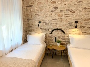 A room with two beds, a stone wall, a small table with a plant, and a window with white curtains. - The Backyard (Eva)