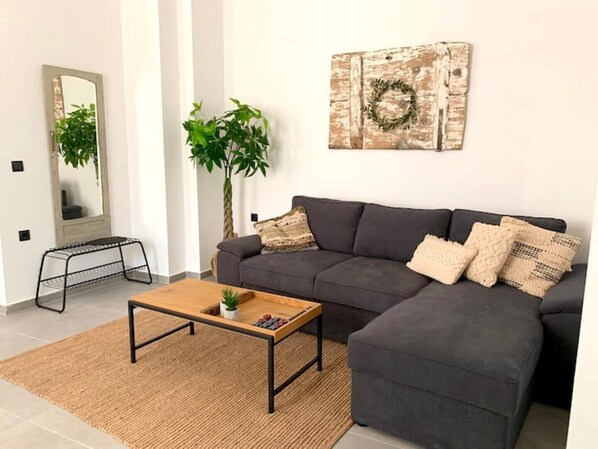 A modern living room with a grey sectional sofa, a wooden coffee table, a small side table, and a large mirror. There is a potted plant and a decorative wreath on the wall. - The Backyard (Eva)