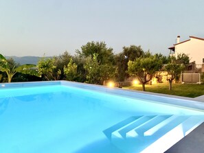 A swimming pool with a clear blue water surface, surrounded by lush greenery and a white building with a red roof in the background.