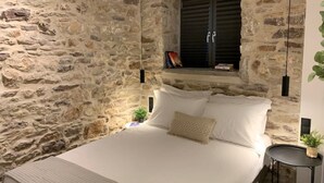 A bedroom with a stone wall, a bed with white linens, a small bedside table, and a window with shutters. - The Backyard (Eva)