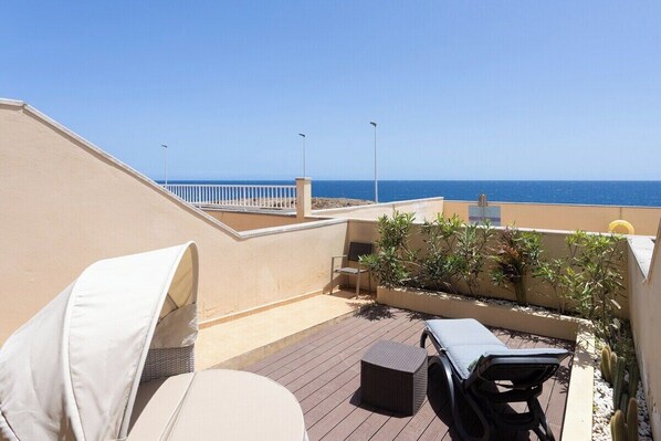 Eine Dachterrasse mit Blick auf das Meer, ein weißer Stuhl, eine Liege und ein kleiner Tisch.