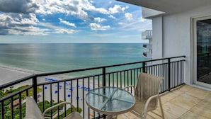 A balcony with a glass table and wicker chairs, overlooking a beach with umbrellas and a clear view of the ocean. - Gorgeous Beachside Condo in superior Longboat Location (Longboat Key)