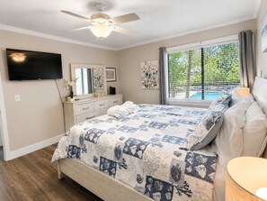 A bedroom with a bed, a ceiling fan, a TV, a dresser, and a window with a view of trees.