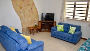A living room with two blue sofas, a flat-screen TV on a wooden stand, a stone wall, and a window with blinds. - Aito 2 in Muri (Muri)