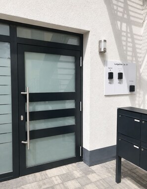 Une porte moderne dotée d’un panneau en verre et d’une poignée en métal, un système d’interphone mural et un meuble de rangement en métal noir.