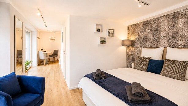 A modern hotel room with a large bed, a blue armchair, a desk with a chair, and a view of the hallway. - Stylish apartment: quiet, 24/7 self check-in & Wi-Fi, street parking (Göttingen)