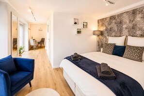 A modern hotel room with a large bed, a blue armchair, a desk with a chair, and a view of the hallway. - Stylish apartment: quiet, 24/7 self check-in & Wi-Fi, street parking (Göttingen)