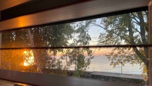 A view through a window with horizontal blinds, a body of water, and trees in the background. - Peaceful Lakefront Living – 3-BR Home with Breathtaking Views; Pet & Smoke-Free (Charter Township of Berlin)