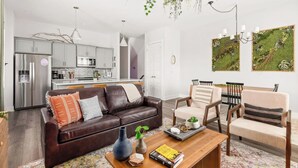 Ein modernes Wohnzimmer mit einer dunkelbraunen Ledersofa, einem Couchtisch mit Büchern und einer Vase, sowie einem Essbereich mit Holzstühlen und einem weißen Tisch. Im Hintergrund ist eine Küche mit Edelstahlgeräten zu sehen.