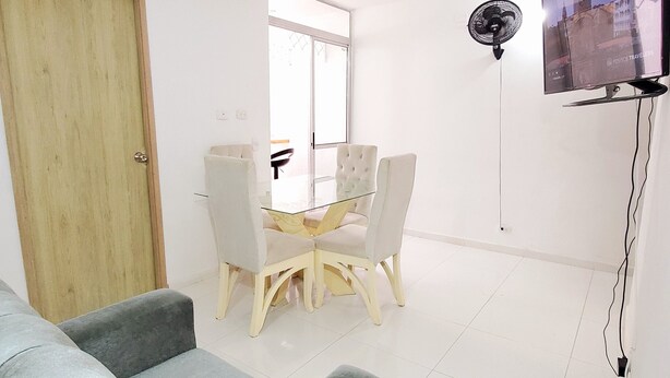 Ein modernes Wohnzimmer mit grauem Sofa, einem Esstisch mit Stühlen, einem an der Wand montierten Fernseher und einer Glastür zum Balkon.
