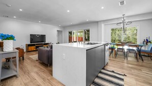 Una cocina moderna con una isla central, un comedor con mesa y sillas, y una sala de estar con sofá y televisión.