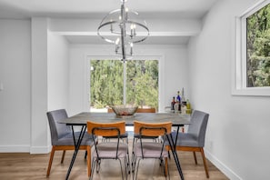 Un coin repas comprenant une table ronde, quatre chaises, un lustre et une vue sur la verdure grâce à de grandes fenêtres.