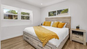 Un dormitorio con estructura de cama de madera, ropa de cama blanca y una manta color mostaza. Hay dos mesitas de noche con lámparas y una planta pequeña en una de ellas.