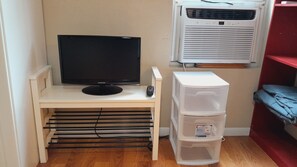 Una habitación pequeña con una televisión sobre un pedestal, un control remoto, un mueble blanco y un estante rojo.