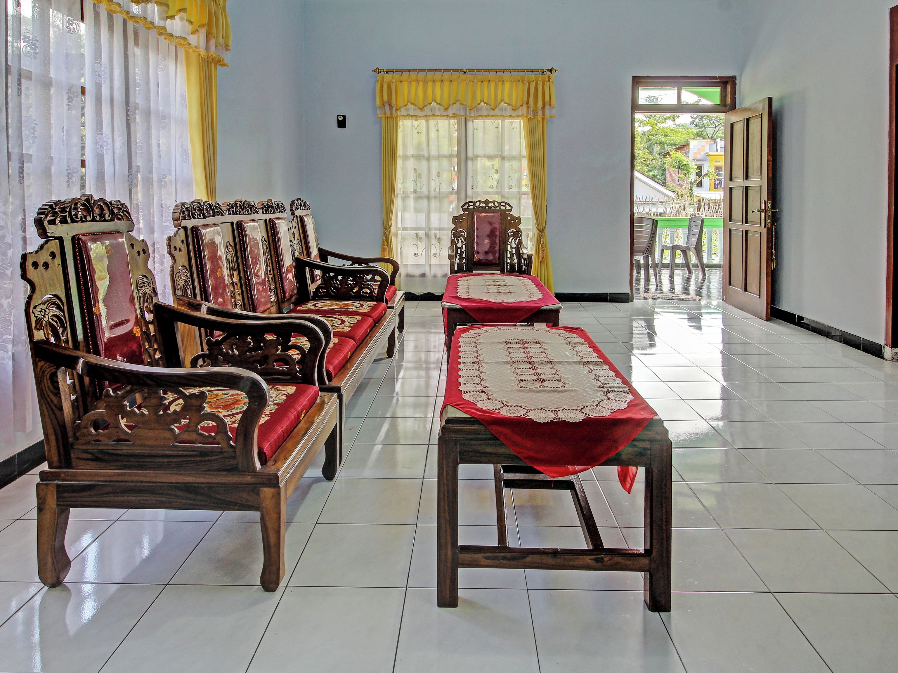 A room with a long wooden bench, two wooden chairs, a small table with a red cloth, and a window with yellow curtains.