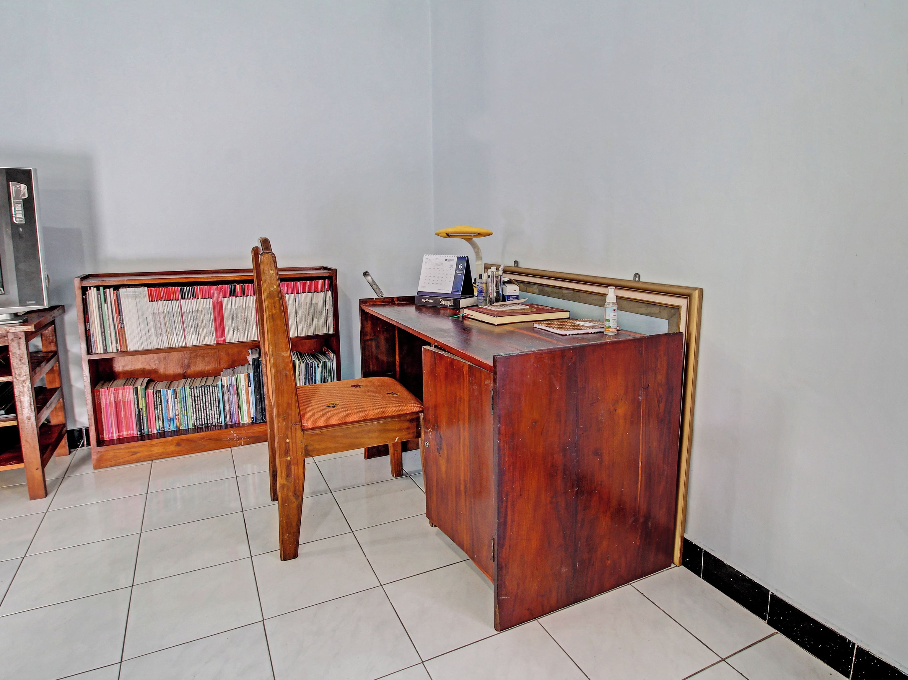 Una habitación con un escritorio de madera, una silla, una estantería y una pequeña mesa con una lámpara.