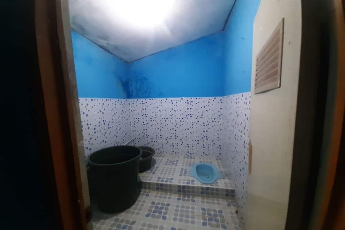 A small bathroom with a blue and white tiled floor, a black bucket, and a blue toilet seat.