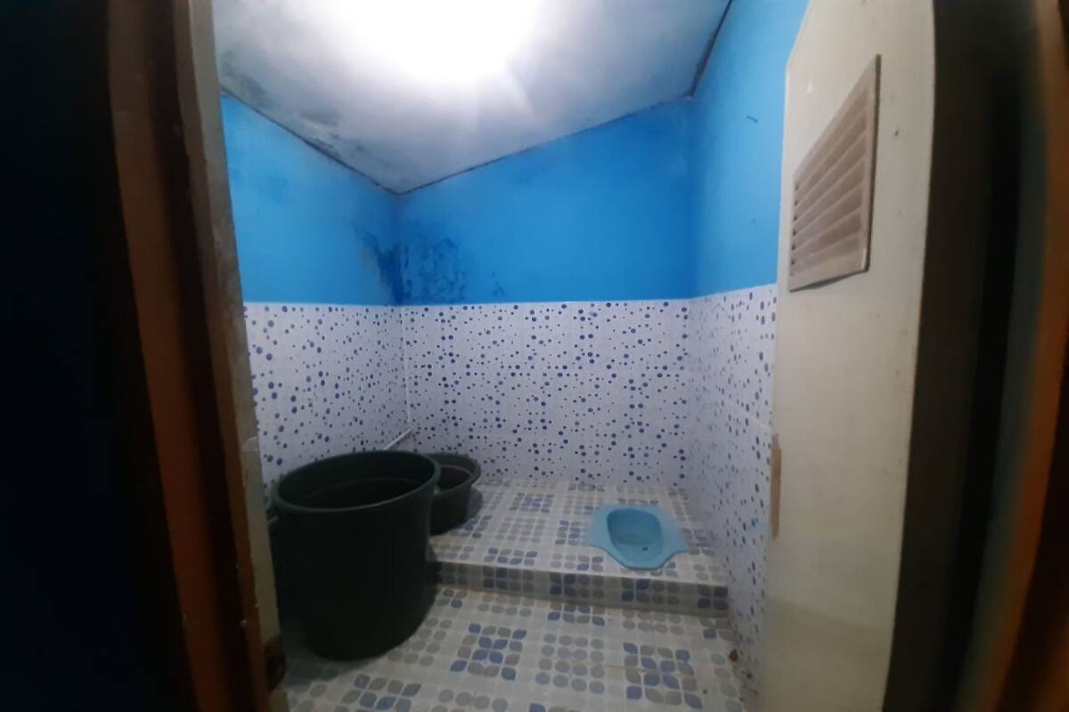 A small bathroom with a blue and white tiled floor, a black bucket, and a blue toilet seat.