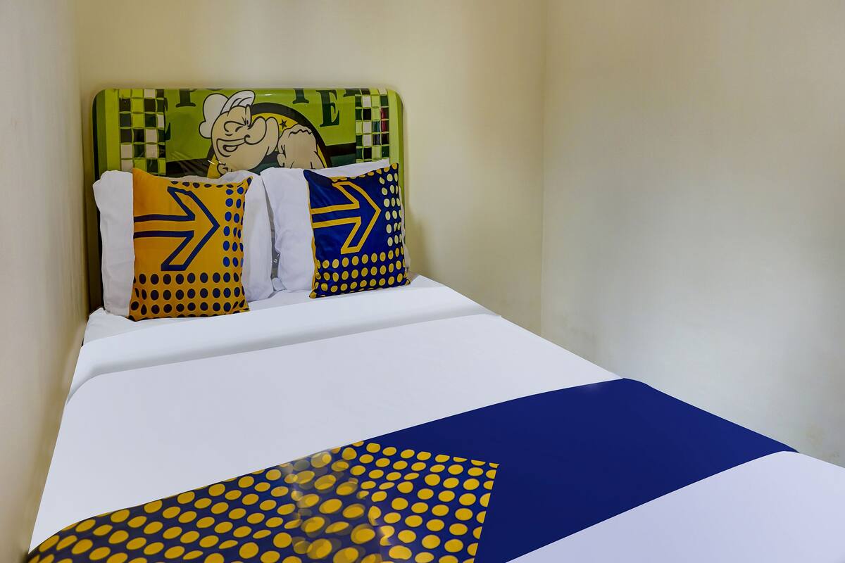Un lit recouvert d’une couvre-lit blanche et bleue, deux oreillers ornés de motifs jaunes et bleus, et une tête de lit verte décorée d’un personnage de dessin animé.