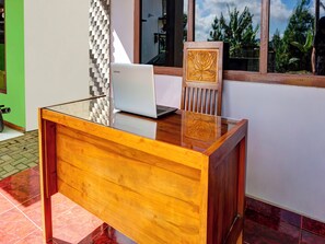A wooden desk with a laptop and a decorative chair, positioned near a window with a view of greenery.