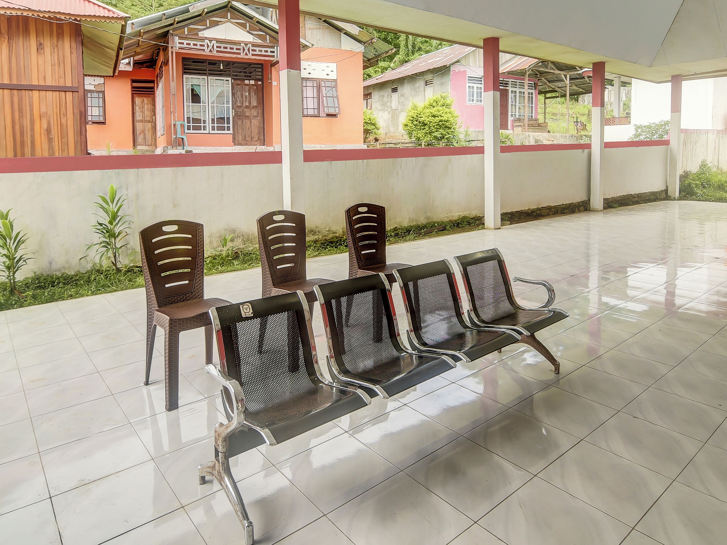 A covered outdoor seating area with chairs and tables, a tiled floor, and a building with a red and white facade in the background.