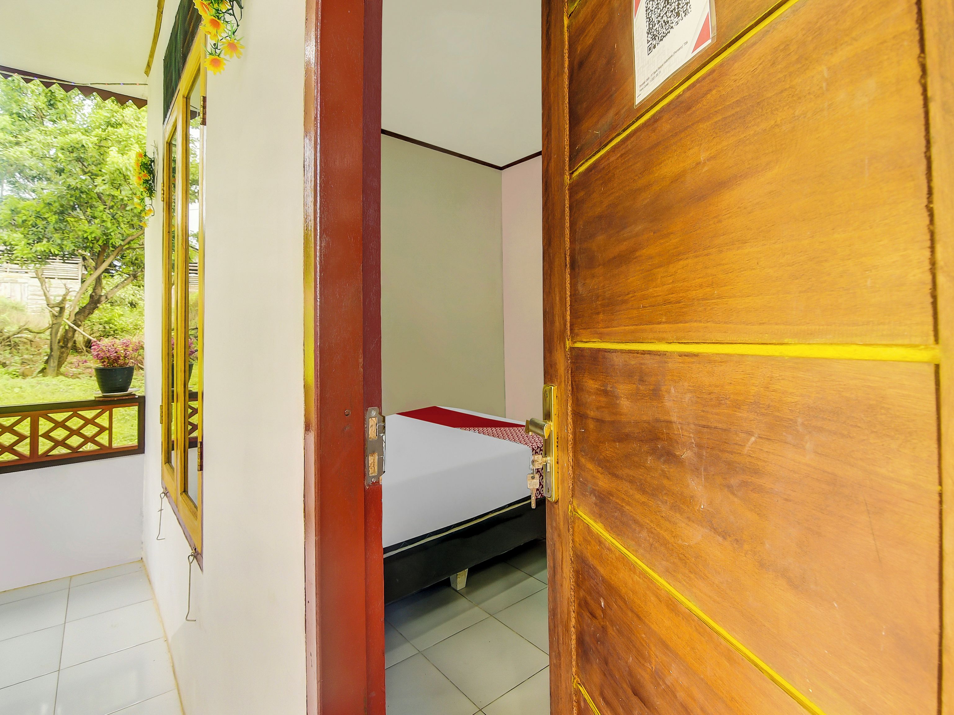 A room with a bed, a window with a view of greenery, and a door with a QR code.